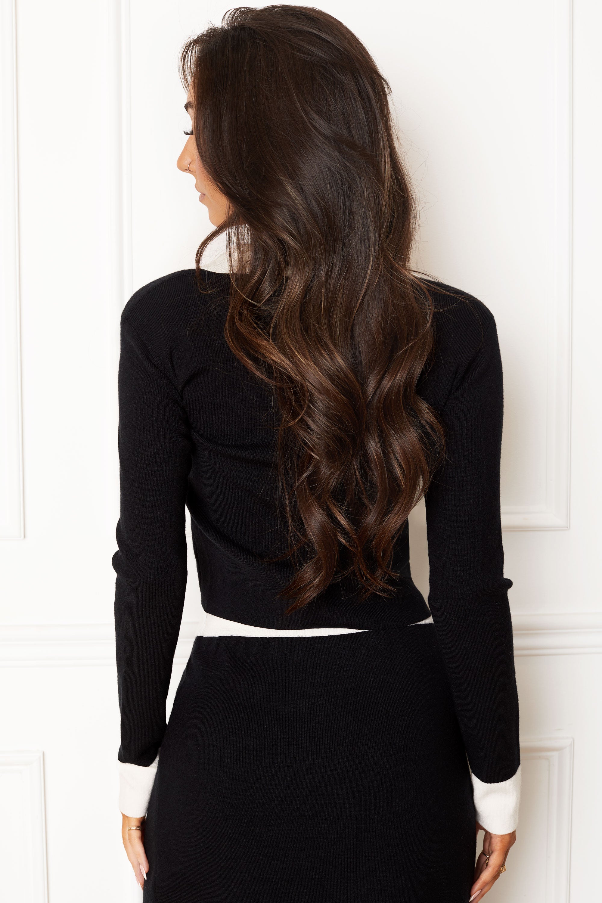 A woman with long, wavy brown hair is shown from behind, wearing a black long-sleeve knit top with contrasting white cuffs and a white waistband, paired with a black skirt. She is standing against a white paneled wall.