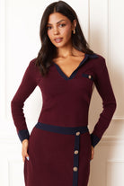 A woman modeling a long-sleeve knit dress in deep burgundy with dark navy blue trim details, including the collar, cuffs, waistband, and front button placket. The dress features a V-neckline, a chest pocket with a decorative button, and a buttoned front detail at the waist.