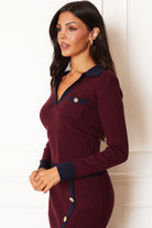 A woman modeling a long-sleeve knit polo dress in deep burgundy with dark navy trim on the collar, cuffs, and button-front hem, posed against a white paneled wall.