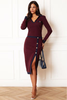 A woman models a long-sleeve burgundy knit midi dress with navy blue trim and silver buttons down the front, holding a navy blue handbag in front of a white paneled wall.