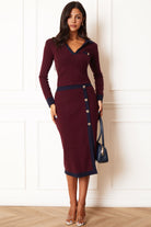 A woman models a long-sleeve burgundy knit top and skirt with navy blue trim and a buttoned side detail, standing against a white paneled wall.