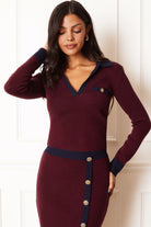 A woman modeling a long-sleeve burgundy knit dress with dark navy blue trim, a matching waist belt with decorative gold-tone buttons, and a vertical button placket down the front.